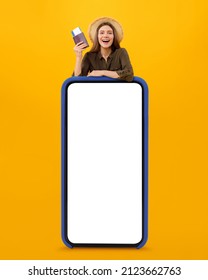 Travel App. Joyful Female Tourist Posing Ner Huge Smartphone With Empty Screen Holding Passport And Boarding Pass Tickets Smiling To Camera Posing Over Yellow Studio Background. Vertical, Mockup