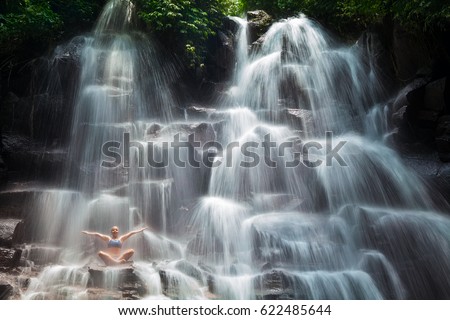 Travel in Bali jungle. Beautiful young woman sitting in yoga pose on rock under falling spring water, enjoy tropic cascade waterfall. Nature day trip, walking adventure, fun on family summer vacation