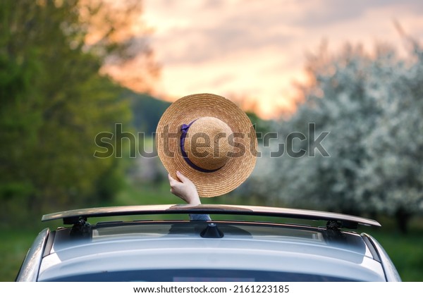 Travel by\
car on vacation. Woman waving with straw hat from car sun roof\
window during sunset. Road trip to\
adventure