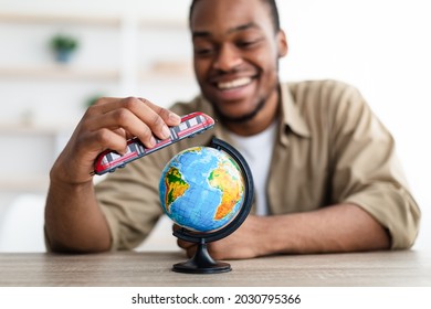 Travel. Cheerful African Guy Playing With Train Model And Globe Sitting At Desk Indoors, Planning His Next Trip. Black Man Dreaming About Journey And Vacation Concept. Selective Focus On Toy