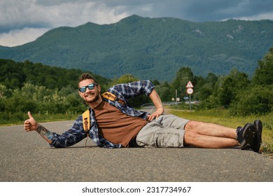 Travel concept. Guy trying to stop car with thumbs up. Young Caucasian man with dreadlocks and beard, hitchhiking in sunglasses and with backpack, lies in the middle of road in mountains.