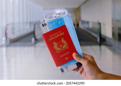 Travel concept: Hand holding SINGAPORE passport, with face mask and boarding pass in airport. Moving walkway travelator in background. Reopening travel lane for safe travels; covid-19 coronavirus.
