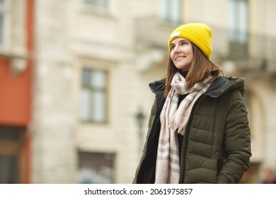 Travel concept: happy smiling young woman posing at street of European city. Model wearing green coat, white and beige scarf, yellow beanie and looking up. Copyspace. Outdoor shot