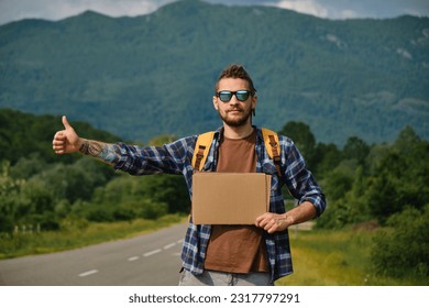 Travel concept. Young man hitchhiker with backpack, stands by mountain country road and holds cardboard sign on sunny summer day. Front view. Guy went on trip alone, trying to stop passing car.