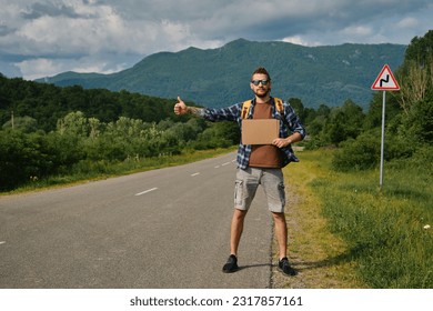 Travel concept. Young man hitchhiker with backpack, stands by mountain country road and holds cardboard sign on sunny summer day. Front view. Guy went on trip alone, trying to stop passing car.