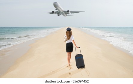 Travel Concept. Young Woman In Flight Attendant Clothes Walking On The Beach With Suitcase And Hat . Overhead Fly Plane. 