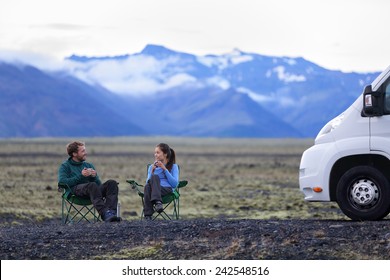 Travel Couple By Mobile Motor Home RV Campervan. People Sitting In Chairs Relaxing Camping And Enjoying Traveling On Iceland In Recreational Vehicle. Young Couple Enjoying Coffee In Nature Landscape.
