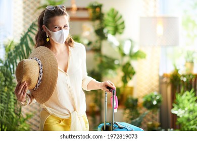 Travel During Covid-19 Pandemic. Stylish Traveller Woman With Medical Mask, Wheel Bag, An Antibacterial Agent, Straw Hat And Sunglasses At Modern Home In Sunny Day.