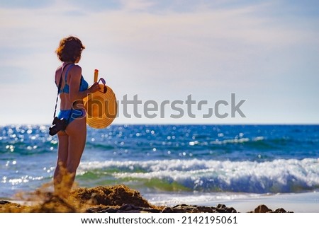 Travel freedom concept. Tourist mature woman in bikini with camera walking on beach, taking photos, sunbathing, enjoying summer vacation.