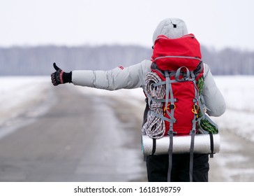 Travel Hitch Hiker Holding Thumb Up On Secondary Road To Share A Ride. Winter Time, Back View, Selected Focus. Big Backpack And Warm Sportawear. Adventure, Independence, Activity, Good Fortune, Risk