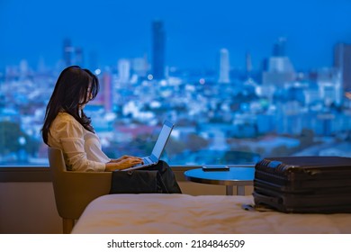 Travel Insurance Concept - Asian Woman Prepare Suitcase For Holiday On Bed. Woman Planning Vacation Trip, Booking Hotel,search Travel Insurance On Laptop, Sitting At Window Enjoying The City At Sunset