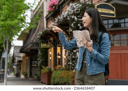 travel lifestyle and people on vacation in usa. cheerful asian female traveler pointing at a landmark in distance with a guidebook in hand on street corner while visiting Carmel by the sea