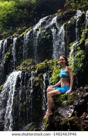 Travel lifestyle. Young traveler woman sitting on the rock, enjoying waterfall landscape. Happy woman wearing blue bikini. Vacation in Asia. Banyu Wana Amertha waterfall Wanagiri, Bali, Indonesia.