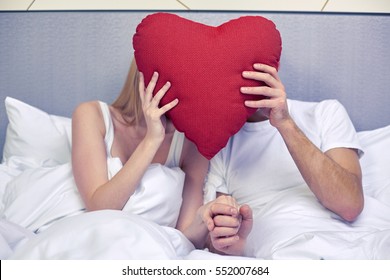 travel, love, valentines day, holidays and happiness concept - happy couple in bed hiding faces behind red heart shape pillow at hotel or home