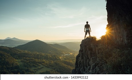 Путешествующий турист один на краю скалы горы и смотрит на долину. Силуэт человека на высокой скале на закате. Пешие походы, приключенческий образ жизни,