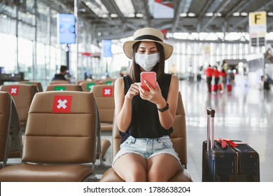 Travel New Normal Under COVID-19 Virus Concept, Happy Traveler Asian Woman With Mask And Luggage Using Mobile Phone And Sit Social Distancing Chair In Terminal Airport, Thailand