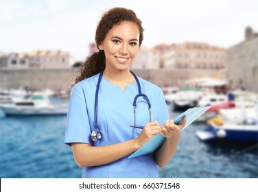 Travel Nursing Concept. Yong Doctor With Clipboard On Cityscape Background