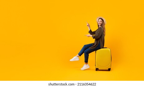 Travel Offer. Happy Female Tourist Pointing Fingers Aside At Free Space For Text Sitting On Suitcase Over Yellow Studio Background, Smiling To Camera. Full Length Shot, Panorama