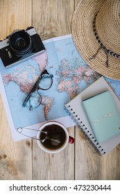 Travel Preparations Essentials. World Map, Cup Of Tea, Vintage Film Camera, Hat, Passport, Notebook And Stylish Glasses On The Old Rustic Wooden Table Background, Flat Lay