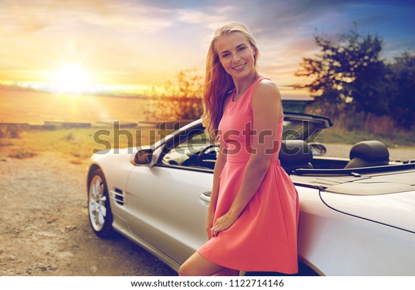 travel, road trip\
and people concept - happy young woman posing at convertible car\
over sunset background