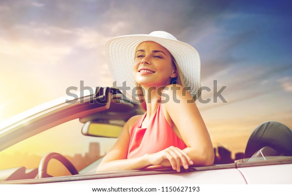 travel, road\
trip and people concept - happy young woman wearing sun hat in\
convertible car over city sunset\
background