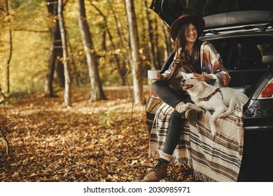 Travel And Road Trip With Pet. Stylish Happy Woman Traveller With Cup And Backpack Sitting With Cute Dog In Car Trunk In Sunny Autumn Woods. Young Female Traveling With Swiss Shepherd White Dog