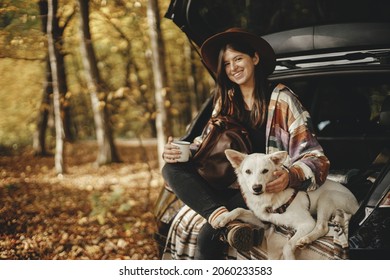 Travel And Road Trip With Pet. Stylish Happy Woman Traveller With Cup And Backpack Sitting With Cute Dog In Car Trunk In Sunny Autumn Woods. Young Female Traveling With Swiss Shepherd White Dog