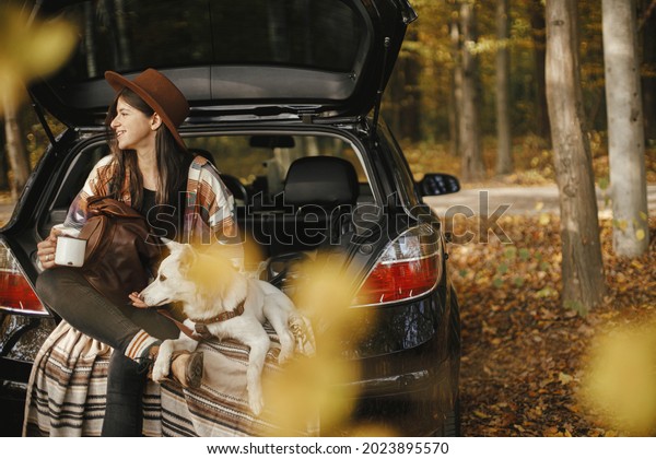 Travel and road trip with pet. Stylish happy woman\
traveller with cup and backpack sitting with cute dog in car trunk\
in sunny autumn woods. Young female traveling with swiss shepherd\
white dog