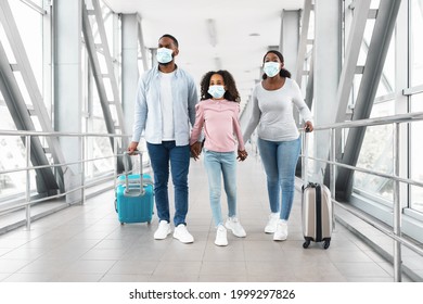 Travel Safe During Coronavirus Pandemic. Portrait of black family of three wearing protective medical masks walking in airport terminal hall, daughter holding hands with mum and dad