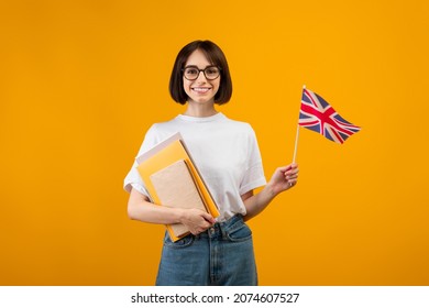 Travel and study, learn English language. Zoomer smiling woman with notebooks and small flag go to university, college or school, isolated on orange background, studio shot - Shutterstock ID 2074607527