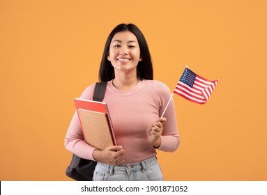 Travel, Study, Student Exchange And Learning English. Zoomer Smiling Asian Woman With Notebooks And Small Flag Of USA Go To American College, Isolated On Beige Background, Studio Shot, Empty Space