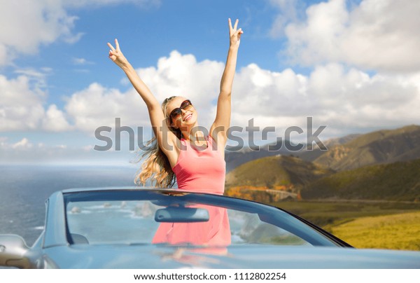 travel, summer holidays, road trip and people\
concept - happy young woman wearing sunglasses in convertible car\
showing peace sign over bixby creek bridge on big sur coast of\
california background