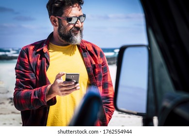 Travel And Technology Concept With Portrait At Handsome Adult Man With Beard Use Modern Phone Outside His Car With Blue Sky And Sandy Beach In Background - Roaming Internet Connection Concept