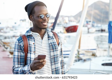 Travel, Tourism, Communication, Technology And People Concept. Handsome Black Man With Backpack Wearing Stylish Hat And Shades Texting Messages On Smart Phone, Enjoying Nice Sunny Weather Outdoors