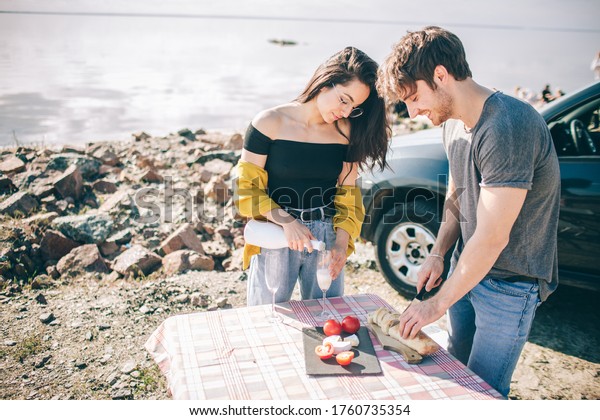 Travel, tourism - Man and woman drink\
champagne near the water at a folding portable table. Picnic near\
the water. Couple going on Adventure. Car travel\
concept.