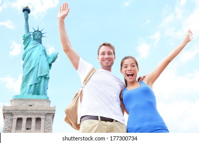 Travel Tourists Couple At Statue Of Liberty, New York City, USA. Multiracial Tourist Couple On Summer Vacation Holidays Cheering Celebrating Happy. Asian Woman, Caucasian Man.