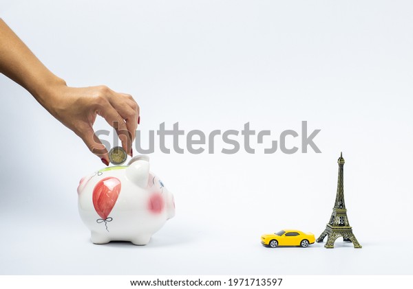 Travel or vacation money saving concept. Hand
hold coin, pink piggy bank with car and Eiffel tower isolated on
white background. Vacation budget saving by working. Europe Holiday
planning concept