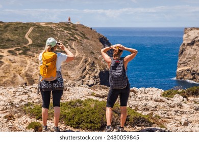 Travel vacation. Two middle age women hiking at trekking trail on seashore in Algarve, Portugal. Hikers looking at lighthouse Cabo de Sao Vicente from coastal Sagres viewpoint - Powered by Shutterstock