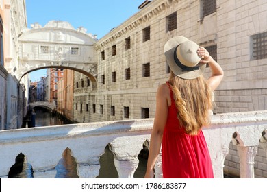 Travel in Venice. Back view of pretty girl in elegant red dress holding hat looking at Bridge of Sighs in Venice, Italy. Beautiful young woman visiting Europe.
