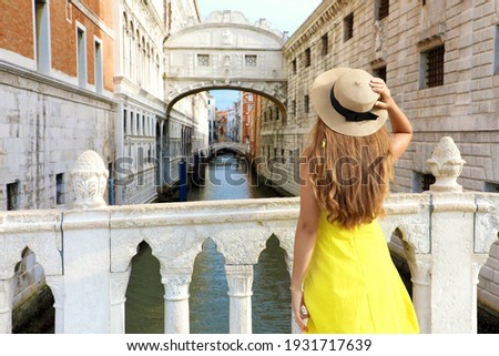 Travel in Venice. Back view of pretty girl in yellow dress holding hat looking at Bridge of Sighs in Venice, Italy. Beautiful young woman visiting Europe.
