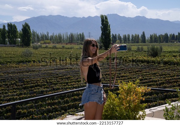 Travel. View of a young woman taking a\
selfie with her cellphone in the vineyard. The grapevines\
plantation and mountains in the background.\
