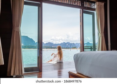 Travel Woman In White Dress Sitting Outdoor Near Wooden House On Water. View On Tourist Woman Relaxing And Leisure From Raft House On Cheow Lan Lake In Thailand. Eco Tourism