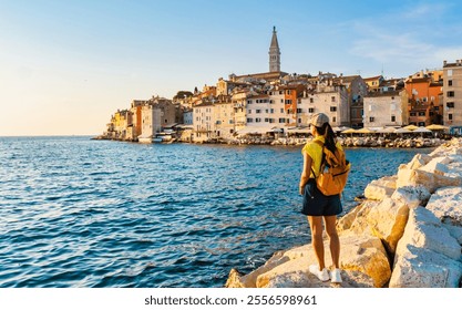 A traveler admires the stunning coastal view of Rovinj, Croatia, as golden sunlight illuminates the historic buildings and calm waters, creating a tranquil atmosphere at dusk sunset - Powered by Shutterstock