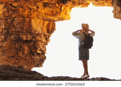 Traveler with a backpack and hat standing at the luminous mouth of a natural sea cave, observing the bright light of the outside world and feeling the spirit of discovery