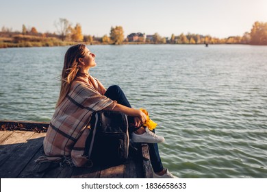 Traveler with backpack relaxing by autumn river at sunset. Young woman sitting on pier breathing free feeling happy