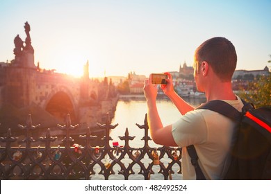 Traveler With Backpack Taking Photo By Mobile Phone. Young Tourist Near Charles Bridge At The Sunset. Prague, Czech Republic. - Back Lit