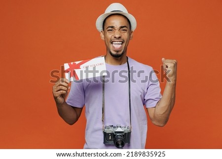 Traveler black man wear purple t-shirt hat hold coupon voucher do winner gesture isolated on plain orange background. Tourist travel abroad on spare time rest getaway. Air flight trip journey concept