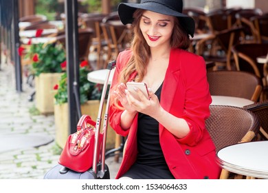 Traveler Buisness Woman Using Cell Phone In Cafe On The Street. Beautiful Girl Wears Black Hat Is Using Smartphone At Cafe Outdoors.