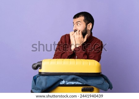 Traveler caucasian man with a suitcase full of clothes over isolated purple background nervous and scared putting hands to mouth