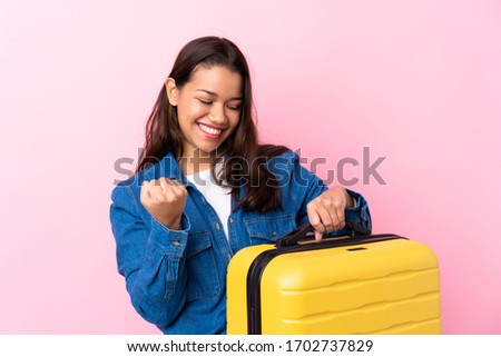 Traveler Colombian woman holding a suitcase over isolated pink background celebrating a victory